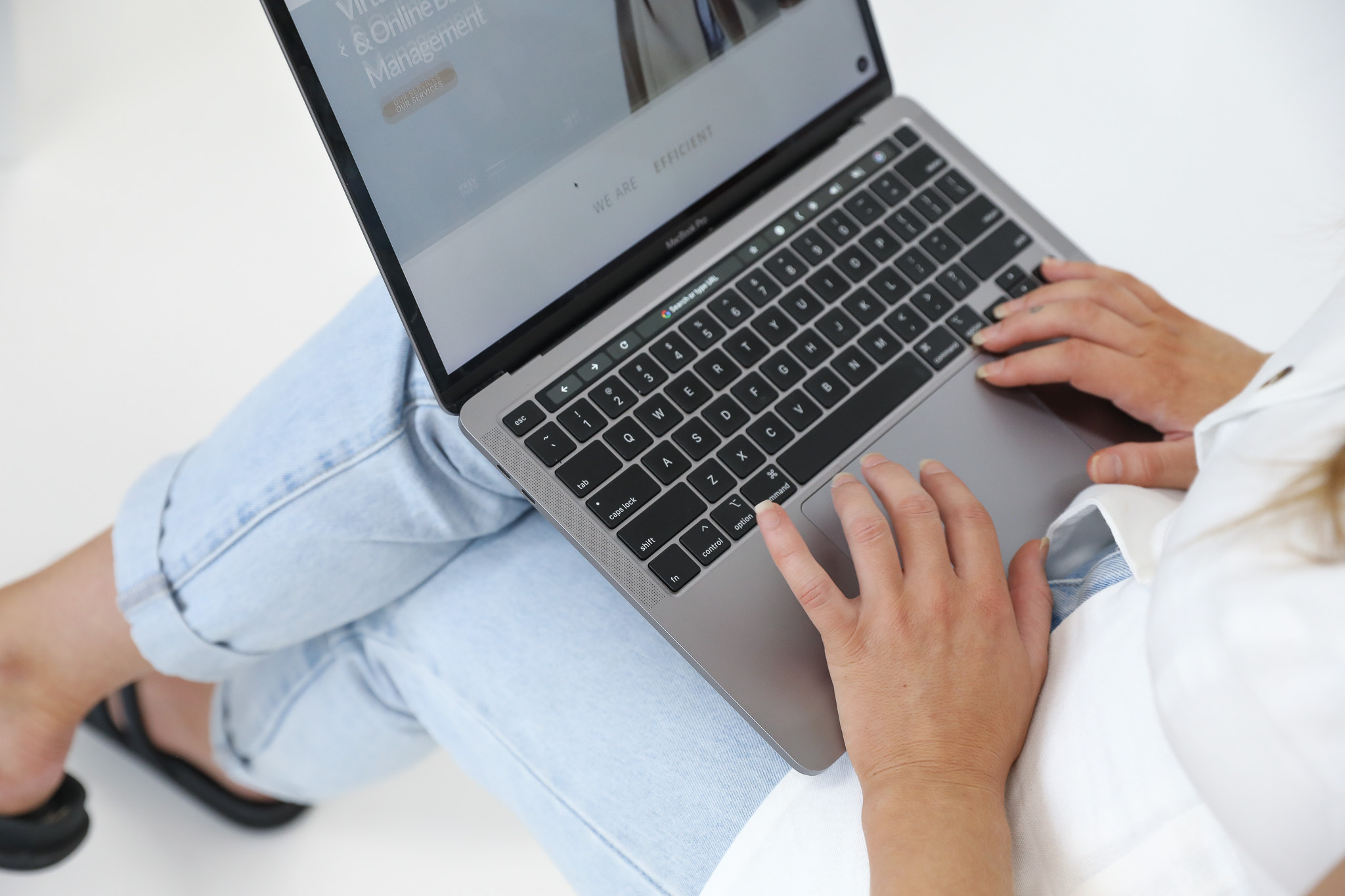 woman in white top, blue jeans and black sandals sitting cross-legged working on laptop on lap